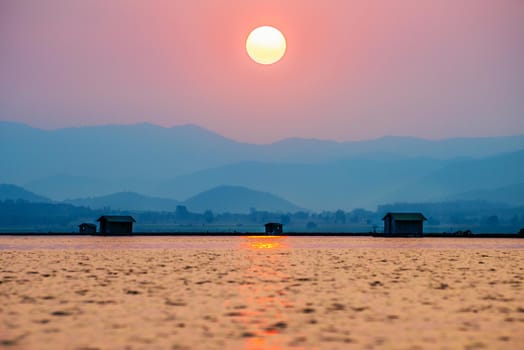 Beautiful nature landscape orange sun in the red sky sunlight over water lake blue mountain background at sunset, silhouette fish farm cages rural lifestyle at Krasiao Dam, Suphan Buri, Thailand