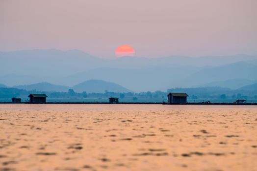 Beautiful nature landscape orange sun in the red sky sunlight over water lake blue mountain background at sunset, silhouette fish farm cages rural lifestyle at Krasiao Dam, Suphan Buri, Thailand