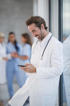 A smiling doctor surfing net on his smartphone while having quick break in a hospital hallway during the Covid-19 pandemic.