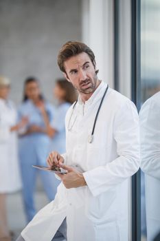 A worried doctor using digital tablet and looking out the window while having quick break in a hospital hallway during the Covid-19 pandemic.