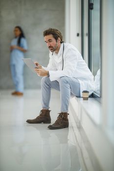 Shot of a mature doctor using digital tablet while sitting near a window in a hospital hallway during the Covid-19 pandemic.