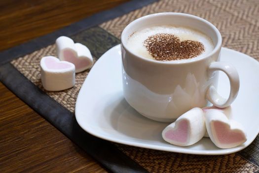 Cup of coffee on a napkin with cocoa heart and sweet marshmallow hearts on a brown table.