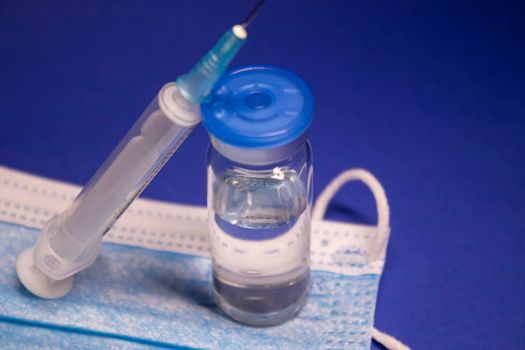 Vaccine bottle phial with no label on blue medical mask and medical syringe with injection needle. isolated on blue background. cure. Development of coronavirus vaccine COVID-19. copy space