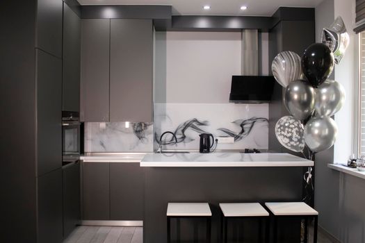 Corner of stylish Minimalistic grey kitchen, dark gray cupboards, white countertops and bar with stools. black sink, kettle and stove. and black and white balloons. High quality photo