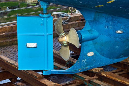 A blue ship in dry dock showing the propeller and rudder steering mechanism from the back