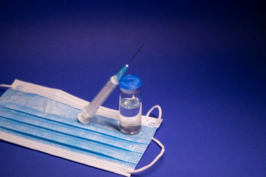 Vaccine bottle phial with no label on blue medical mask and medical syringe with injection needle. isolated on blue background. cure. Development of coronavirus vaccine COVID-19. copy space