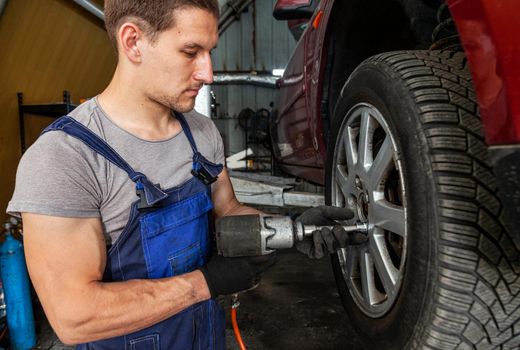 A muscular mechanic is maintaining wheels on a vehicle in auto garage with pneumatic tool.