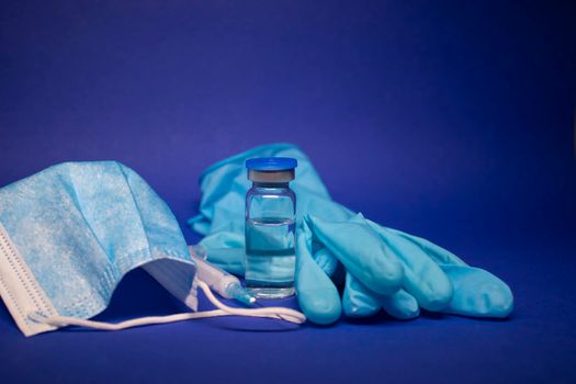 Vaccine bottle phial with no label, medical syringe with injection needle, blue medical mask and gloves . isolated on blue background. cure. Development of coronavirus vaccine COVID-19. copy space