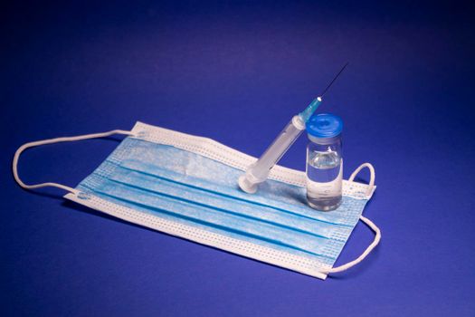 Vaccine bottle phial with no label on blue medical mask and medical syringe with injection needle. isolated on blue background. cure. Development of coronavirus vaccine COVID-19. copy space