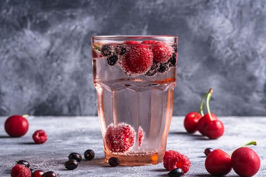 Fresh cold sparkling water drink with cherry, raspberry and currant berries in red faceted glass on stone concrete background, summer diet beverage, angle view