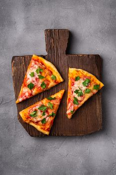 Hot pizza slices with mozzarella cheese, ham, tomato and parsley on brown wooden cutting board, stone concrete background, top view