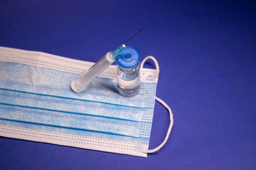 Vaccine bottle phial with no label on blue medical mask and medical syringe with injection needle. isolated on blue background. cure. Development of coronavirus vaccine COVID-19. copy space