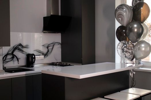 Corner of stylish Minimalistic grey kitchen, dark gray cupboards, white countertops and bar with stools. black sink, kettle and stove. and black and white balloons. High quality photo