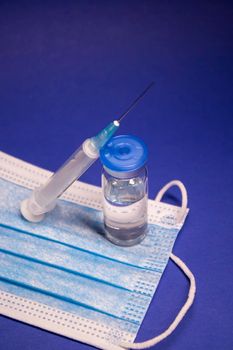 Vaccine bottle phial with no label on blue medical mask and medical syringe with injection needle. isolated on blue background. cure. Development of coronavirus vaccine COVID-19. copy space