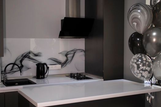 Corner of stylish Minimalistic grey kitchen, dark gray cupboards, white countertops and bar with stools. black sink, kettle and stove. and black and white balloons. High quality photo