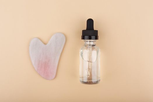 Top view of skin serum in transparent bottle with black cap and pink heart shaped guasha massager against light beige background