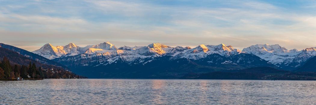 Stunning panorama view of famous Swiss Alps peaks on Bernese Oberland Eiger North Face, Monch, Jungfrau at dusk from Lake Thun (Thunersee) on a sunny autumn day with blue sky cloud,Bern, Switzerland