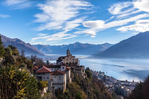 Stunning aerial panorama view of Madonna del Sasso church above Locarno city with Lake Maggiore, snow covered Swiss Alps mountain peaks and blue sky cloud in background in autumn, Ticino, Switzerland