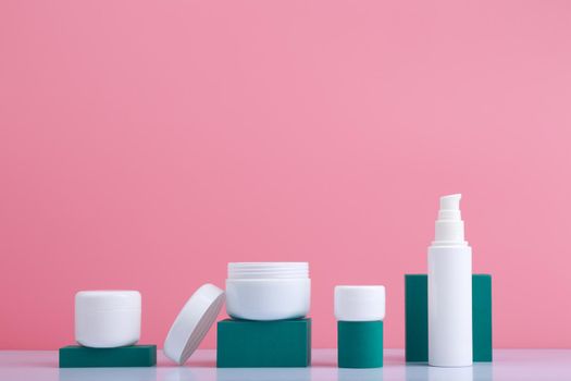 Set of cosmetic bottles on white table with green geometric podiums against pink background with space for text. Concept of skin care cosmetic set and beauty treatment