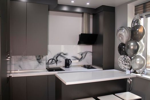 Corner of stylish Minimalistic grey kitchen, dark gray cupboards, white countertops and bar with stools. black sink, kettle and stove. and black and white balloons. High quality photo