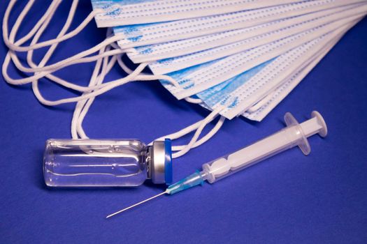 Vaccine bottle phial with no label and medical syringe with injection needle near pile of blue medical masks . isolated on blue background. cure. Development of coronavirus vaccine COVID19. copy space