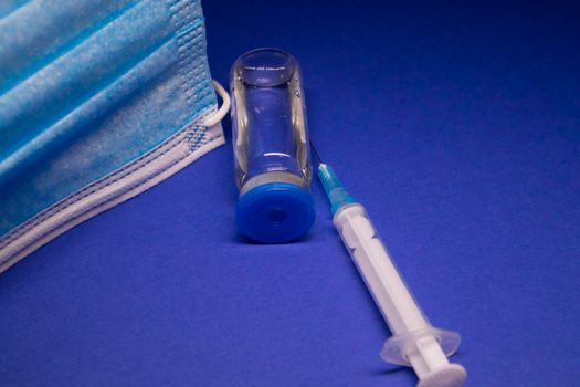 Vaccine bottle phial with no label near blue medical mask and medical syringe with injection needle. isolated on blue background. cure. Development of coronavirus vaccine COVID-19. copy space