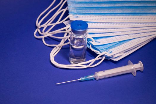 Vaccine bottle phial with no label and medical syringe with injection needle near pile of blue medical masks . isolated on blue background. cure. Development of coronavirus vaccine COVID19. copy space