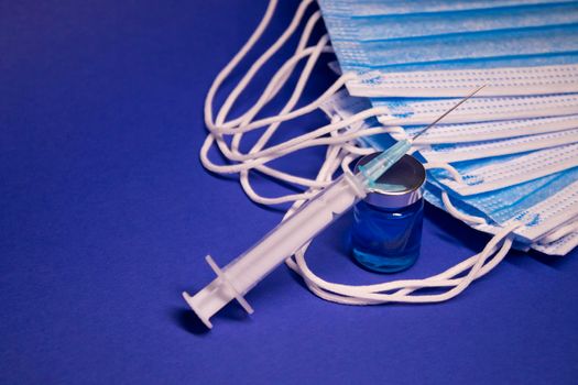 Vaccine bottle phial with no label and medical syringe with injection needle near pile of blue medical masks . isolated on blue background. cure. Development of coronavirus vaccine COVID19. copy space