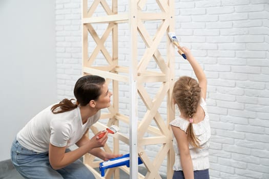 Close up young mother and child holding brush and painting rack in white color in bright room. Concept of repair at home.