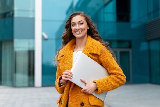 Business woman with laptop dressed yellow coat standing outdoors corporative building background Caucasian female business person on city street near office building Stylish businesswoman
