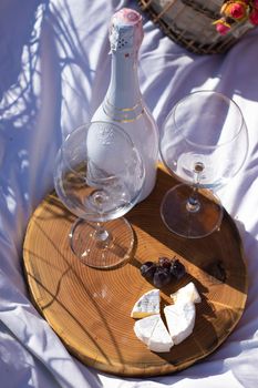 wooden stand with two glasses of champagne and a bottle, grapes and camembert cheese on a white blanket in the field. picnic.
