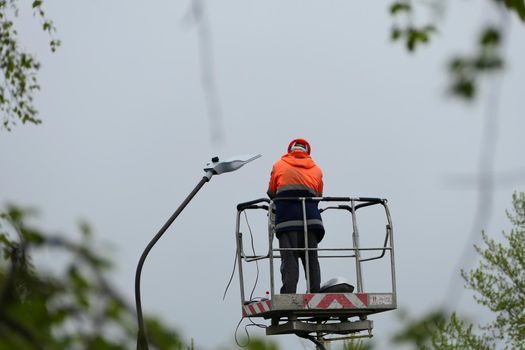 An electrician in special clothing and a helmet repairs a street lamp and screws a street light bulb on a special lifting platform. High quality photo