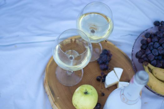 wooden stand with two glasses of champagne and a bottle, grapes and camembert cheese on a white blanket in the field. picnic