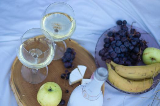 wooden stand with two glasses of champagne and a bottle, grapes and camembert cheese on a white blanket in the field. picnic