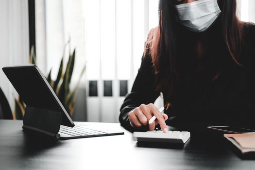businesswoman wear mask working on desk office with using a calculator to calculate the numbers, finance accounting concept