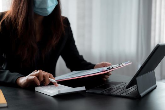 businesswoman wear mask working on desk office with using a calculator to calculate the numbers, finance accounting concept