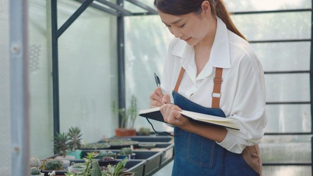 Asian beautiful young gardener woman confident with apron use paper book to checking list growth at greenhouse cactus plant nursery, florist with small business working concept