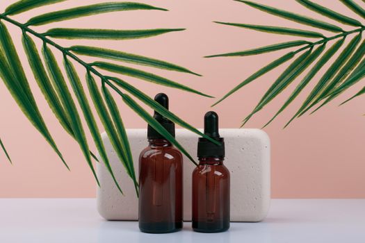 Two glass dark brown cosmetic bottles with skin serum, elixir or oil next to beige rectangular podium against pink background with palm leaves