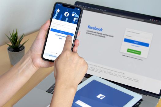 Calgary, Alberta. Canada. Nov 11 2020: A person using Facebook app on an iPhone Pro 12, iPad and a Macbook Pro on a wooden table.