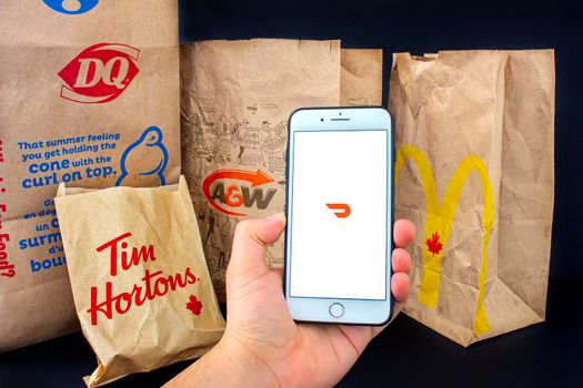 Calgary, Alberta. Canada. April 24, 2020: A person holding an iPhone Plus with the Door Dash application open with delivered food bags from Tim Hourtons, A&W, McDonals and DQ