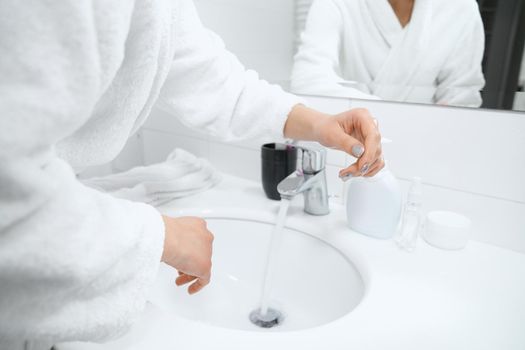 Side view of young woman in white robe standing near sink and washing hands with special antibacterial soap at home. Concept of body procedure for improvements skin.