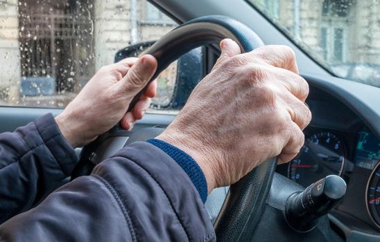 Close-up of the hand of an adult man in a passenger car, holding the steering wheel in cold rainy weather in the city. The man is driving the car.