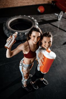 Two young smiling female friends resting after training at the gym. They are holding energy drinks and looking at camera. Selective focus. Top view.