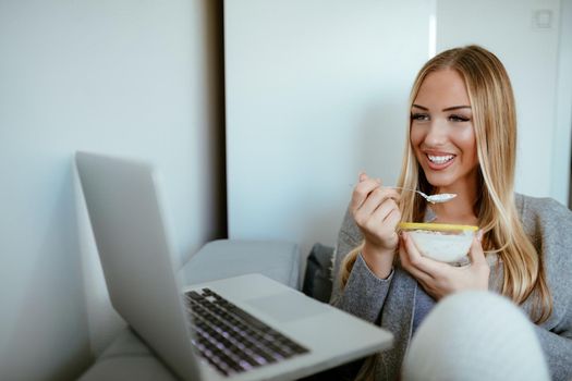 Attractive smiling girl sitting on bed having breakfast and using laptop. She is surfing on the net and checking social media. 