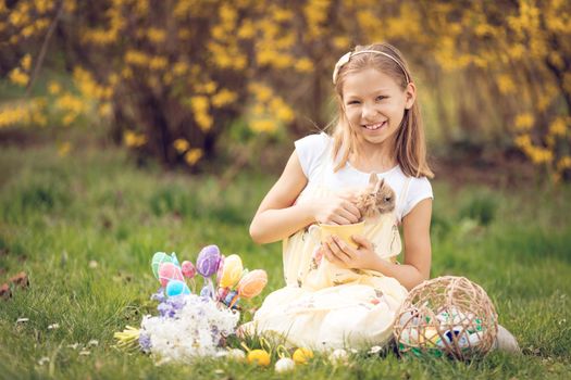 Beautiful smiling little girl holding cute bunny and sitting on the grass with Easter eggs and flowers in spring holidays. Looking at camera. 