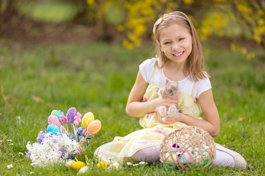 Beautiful smiling little girl holding cute bunny and sitting on the grass with Easter eggs and flowers in spring holidays. Looking at camera.