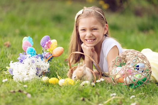 Beautiful smiling little girl lying down in the grass beside cute bunny, Easter eggs and flowers in spring holidays. Looking at camera.