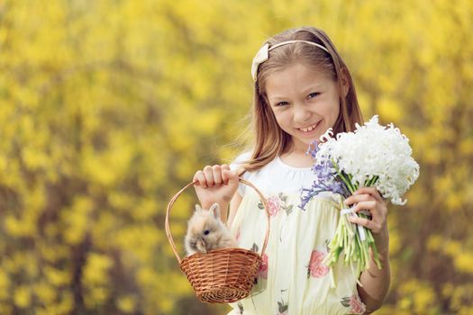 Beautiful smiling little girl holding little basket with cute Easter bunny and bouquet of flowers hyacinth in spring holidays. Looking at camera.