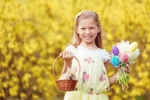 Beautiful smiling little girl holding little basket with cute Easter bunny and bouquet of flowers hyacinth and colorful eggs in spring holidays. Looking at camera. In the background is a many yellow flowers.