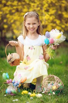 Beautiful smiling little girl holding cute bunny in the basket and bouquet of white flowers and Easter eggs in spring holidays. Looking at camera.